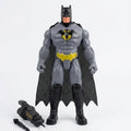 A Batman action figure standing against a plain light gray background, with a black cape, gray suit, yellow chest emblem, and black boots. Two small accessory pieces are placed on the floor to the left.