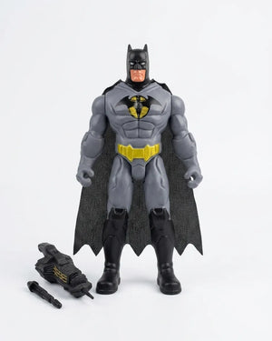 A Batman action figure standing against a plain light gray background, with a black cape, gray suit, yellow chest emblem, and black boots. Two small accessory pieces are placed on the floor to the left.