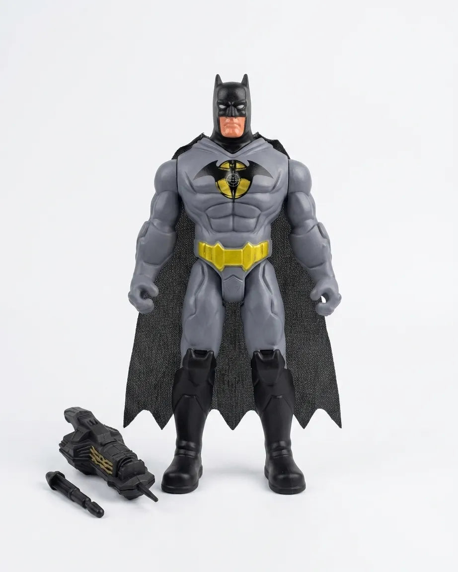 A Batman action figure standing against a plain light gray background, with a black cape, gray suit, yellow chest emblem, and black boots. Two small accessory pieces are placed on the floor to the left.