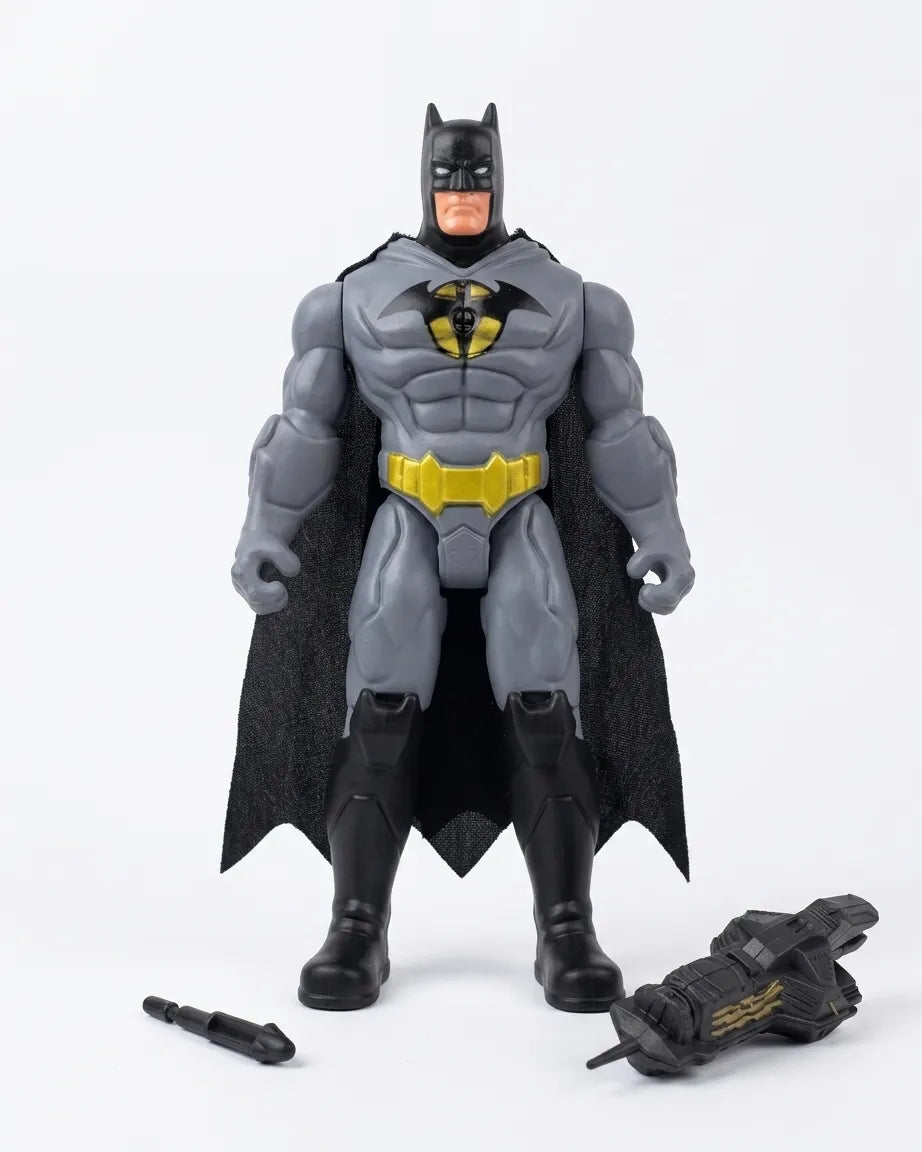 A Batman action figure standing against a plain white background, with a black cape, gray bodysuit, yellow belt and chest emblem, and a small black accessory placed near the base.