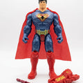 A Superman action figure standing against a white studio background, with a red cape, blue bodysuit, gold belt, and a red weapon accessory placed in front of the figure.