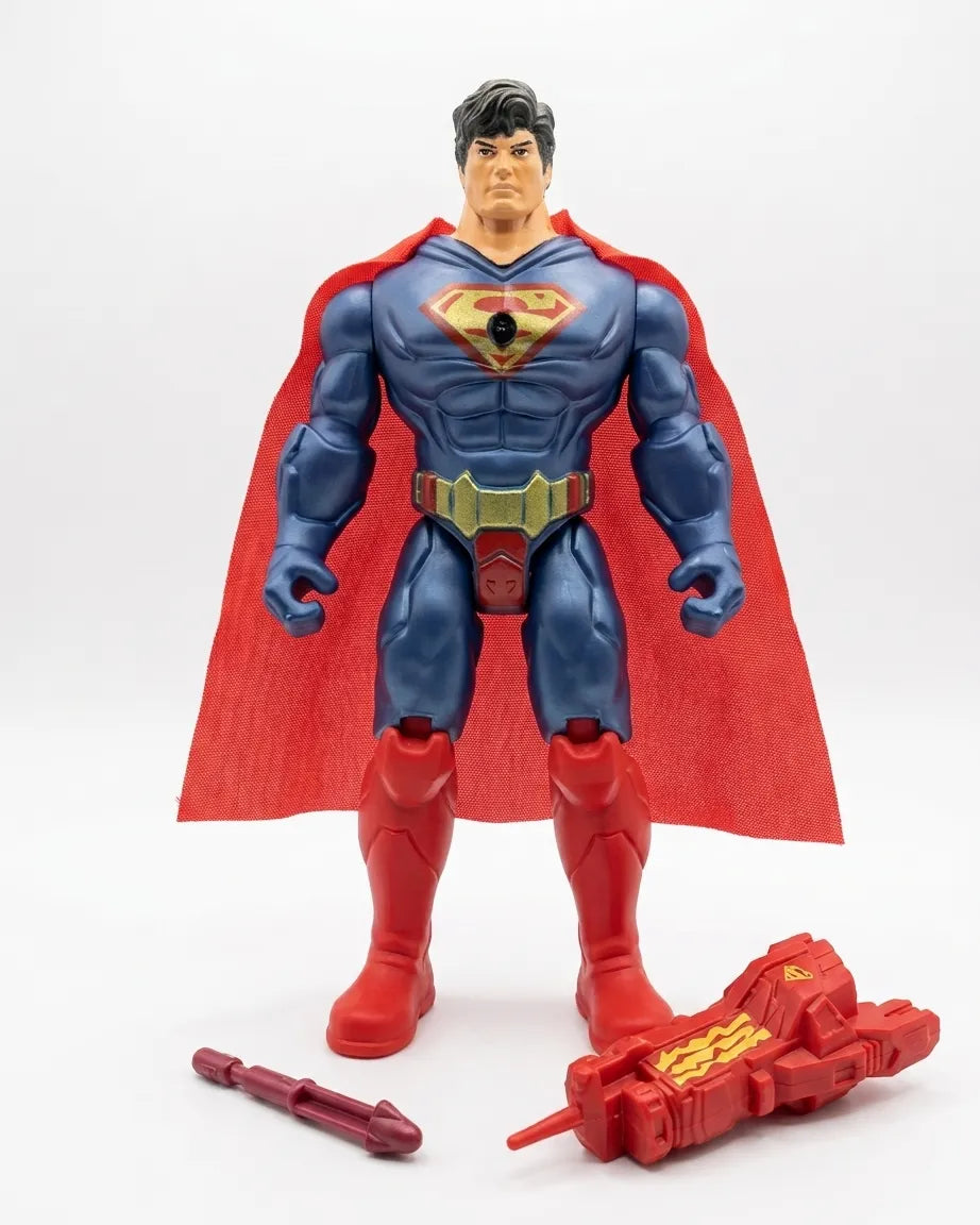 A Superman action figure standing against a white studio background, with a red cape, blue bodysuit, gold belt, and a red weapon accessory placed in front of the figure.