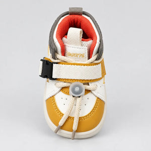 A baby's first shoe featuring a white and mustard yellow color-blocked design, with beige laces tied in a bow, a hook-and-loop strap with a black plastic buckle, and a soft orange inner lining. The brand name 'PORTS' is visible on the tongue label.