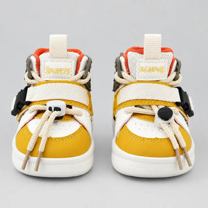 A pair of toddler high-top sneakers shown from the front on a light gray background. The shoes feature a white and mustard yellow color scheme with orange accents, beige laces, and black plastic buckle details.